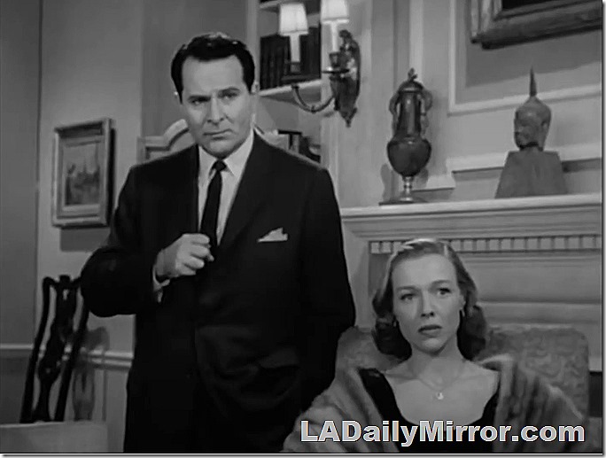Two mystery guests. The man is in a suit with a skinny tie. The woman is seated and is wearing a fur coat. There is bric a brack on the fireplace mantle. The somewhat harsh TV lighting is unmistakable 