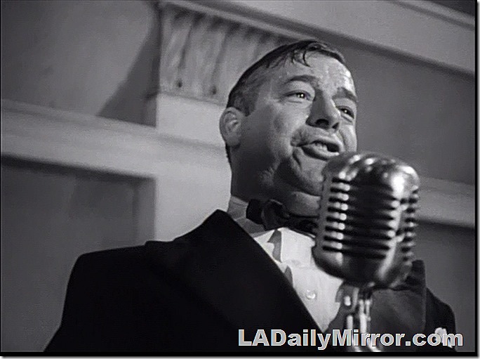 Man in a coat and bow tie speaking into a microphone. 