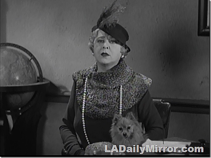 Elegantly dressed woman. She's wearing a hat and a dress with a large collar and a long necklace. She holds a tiny dog. 