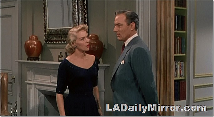 Mystery leading man and mysterious companion stand in front of fireplace. He is neatly dressed in a suit coat and tie and she is wearing a casually elegant black dress with a low neckline. He seems reflective and she is upset. 