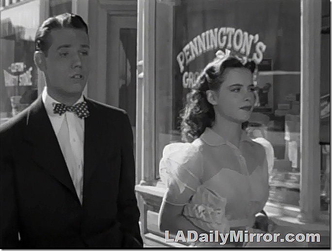 Tall man in a coat, white shirt and bow tie walking with a young woman. 