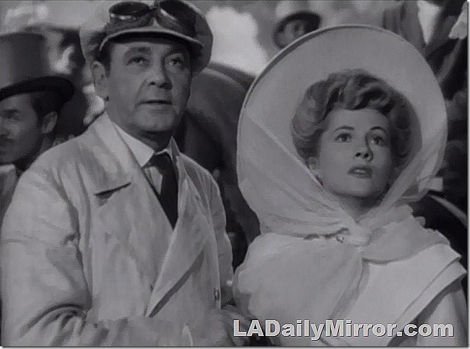 Man in a motoring outfit of the early 20th century, goggles up on his motoring hat. An elegant leading lady in a large hat. They are looking skyward and do not approve of such goings-on. 