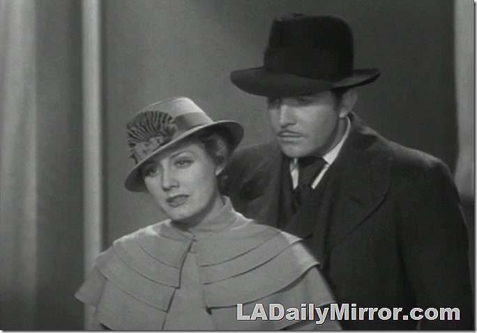 Man and woman. She is elegantly dressed with a cape and hat. He wears a fedora, suit and overcoat. 