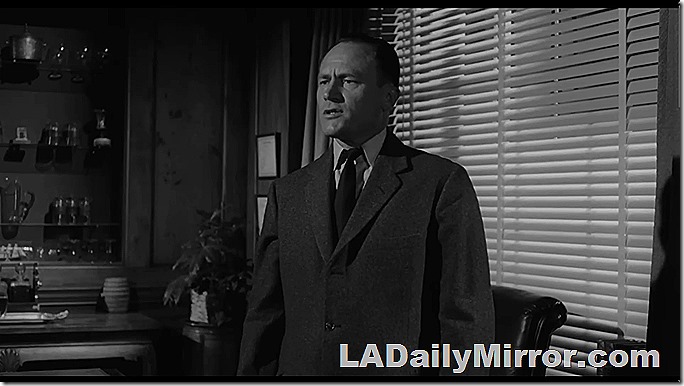 Man in suit and tie stands in front of venetian blinds. Must be a film noir. 