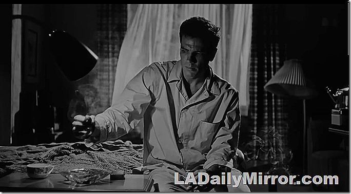 Man in pajamas sitting on bed in funky apartment. Smoking cigarette. 