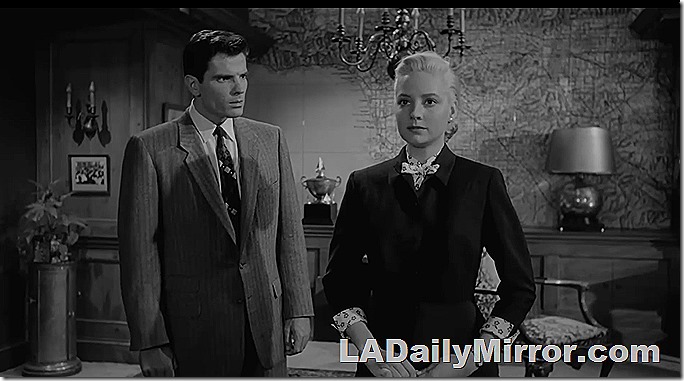 Man in suit looks surprised at blonde woman in suit, staring straight ahead. Also a huge map of L.A. in the background