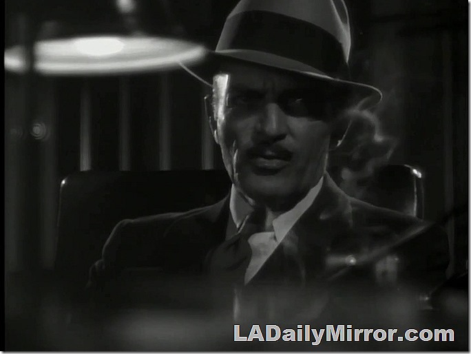Man in suitcoat, tie and hat. Small mustache. The image is dark and moody, with cigarette smoke. 