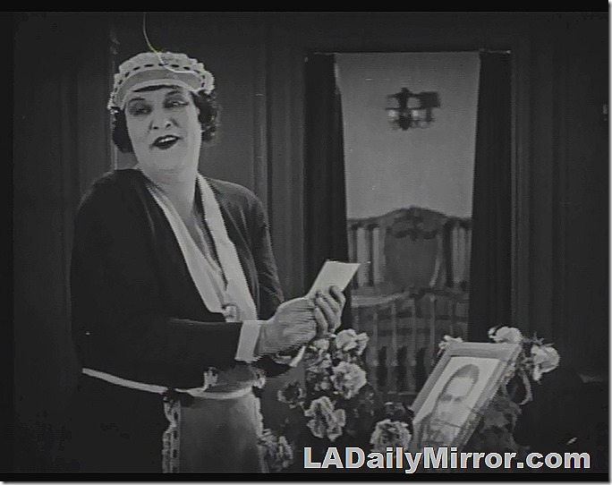 Interior of home. Woman in maid's outfit. Holding a piece of paper. Flowers on table.  Bed is visible through a doorway. 