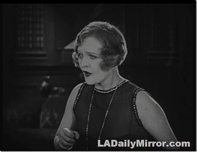 Woman with short hair, sleeveless shift, long necklace. Looks alarmed. 
