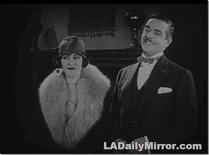 Man in a wing collor and bow tie, with vest and jacket. Woman with fur-trimmed coat, cloche hat