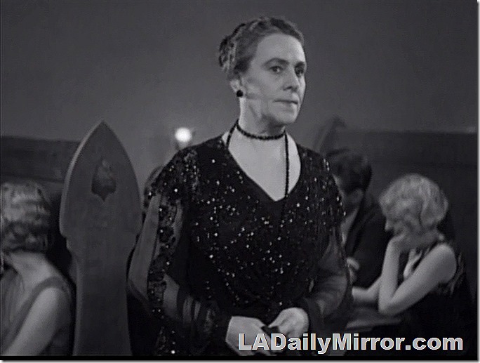 Woman with a short, severe hairstyle and a disapproving expression. She is finely dressed. 