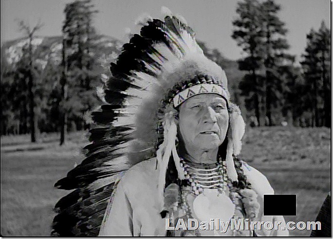 Outdoors. Mountains and trees in background. Man in Plains Indian bonnet. 