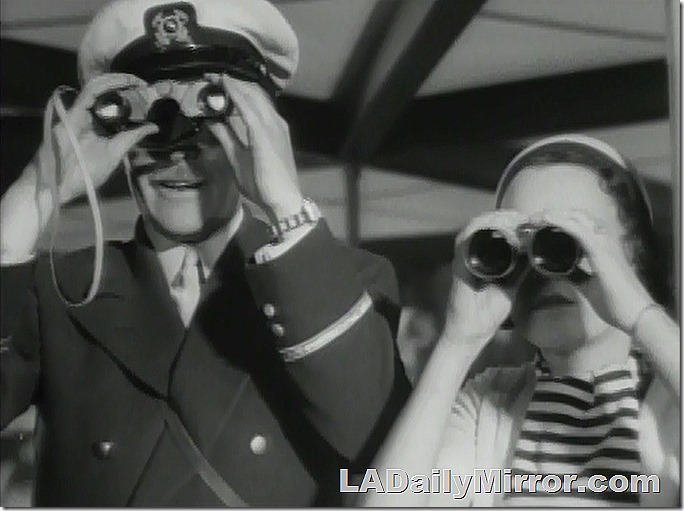 A man in a boater's cap looks through binoculars. A woman next to him also looks through binoculars. A man in a boater's cap looks through binoculars. A woman next to him also looks through binoculars.