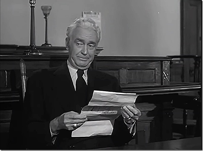 Man with white hair, in coat and tie. He is holding a document. 
