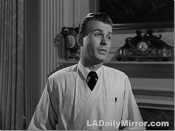 Actor in doctor's smock. On the mantel in the background are a vase and a clock.
