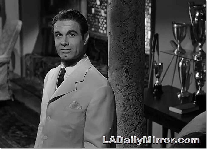 Man with dimpled chin in light colored coat and dark tie. Many trophies are in the background. 