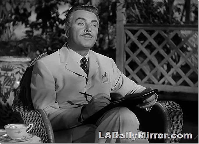 Man with neat mustache wears white suit. He's sitting in a wicker chair, making notes with a pen. 