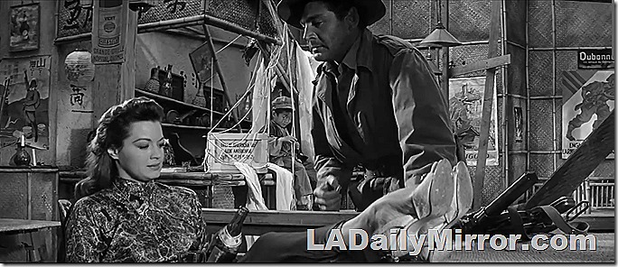 Mystery woman holds bottle. Has feet on table. Mystery leading man is looking at her. 