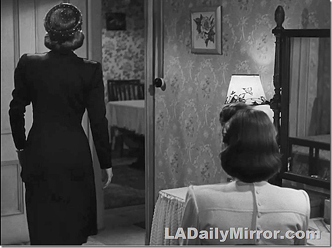 Two Back of the Head Women. One is standing. She's in a dark dress and one shoulder is higher than the other. The second woman is sitting and is wearing a light-colored blouse. 