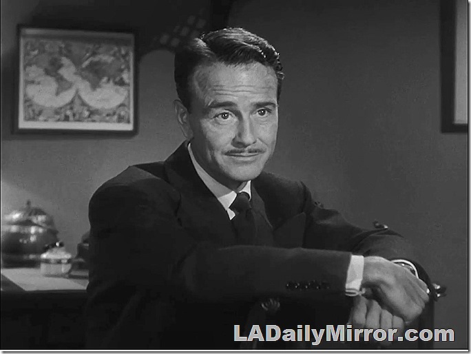 Man with slight mustache. Arms folded over back of chair.