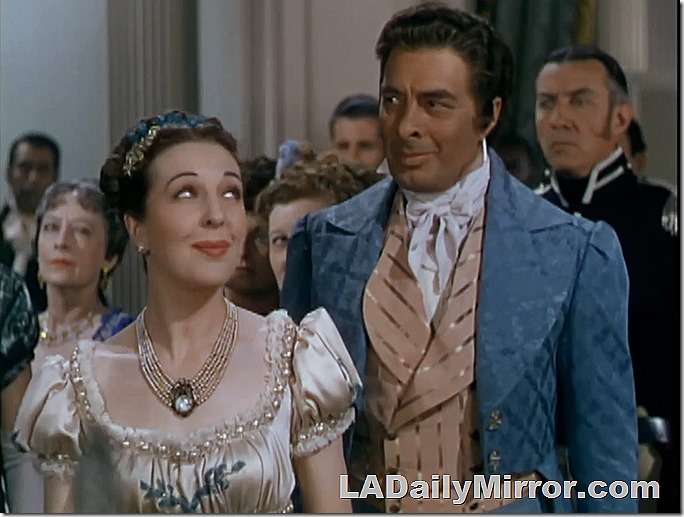 Woman in elegant dress with elaborate necklace. Man in coat with wide lapels and ornate vest. 