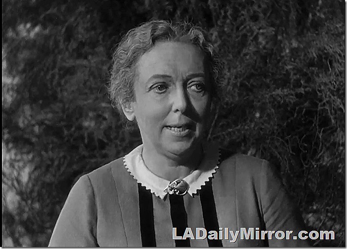 Older in a dress with a narrow, scalloped collar and a brooch at her neck. 