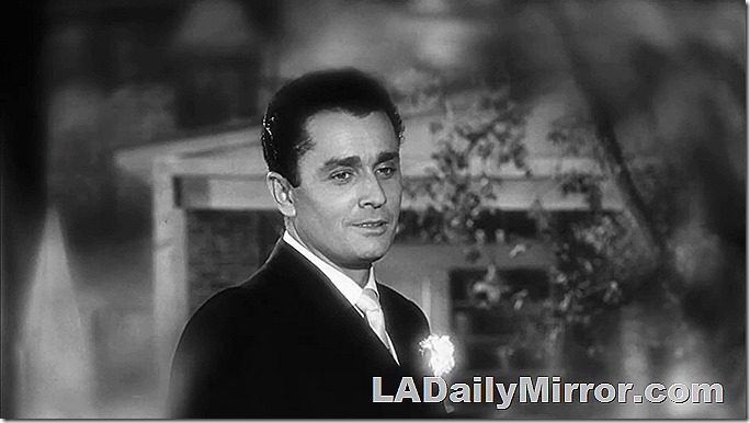 Man in suit, with flower in lapel. The background is blurry. 