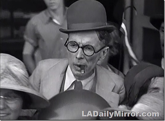 Man in coat and derby, wearing glasses and smoking a cigar. 