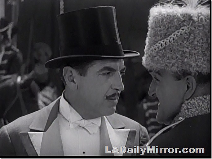 Man with small mustache, upturned at the ends. He's in a top hat and a most impressive coat. His companion is wearing a furry hat. They are up to no good, I think. 