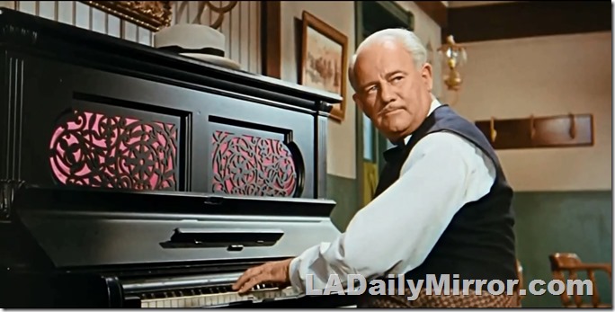Older man in vest, playing upright piano. 