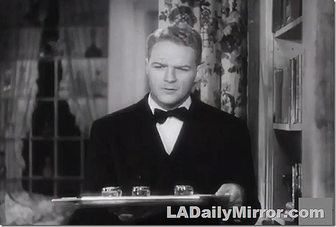 Man in dark coat and bow tie, holding a tray of glasses. He has a quizzical expression. 