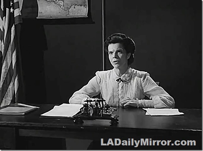 Woman sitting in front of a blackboard. An American flag is in the corner and a map is hanging in the background. She has papers on her desk. 