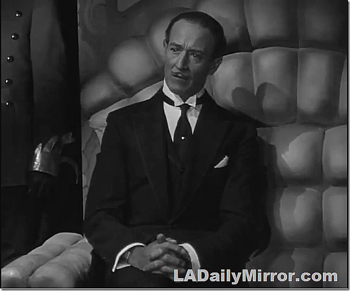 Man with slight mustache, in coat, tie and wing collar. He is sitting with his hands folded. 