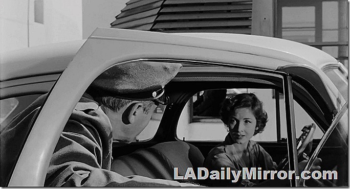 Woman at the steering wheel of a car. Back of the Head Guy has the door open and is talking to her. 