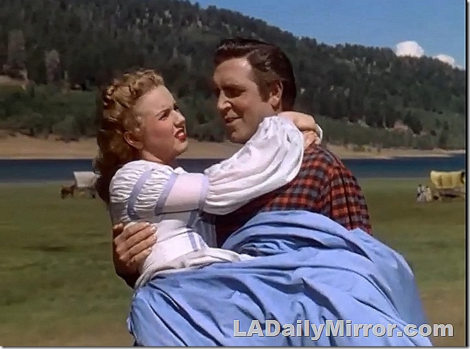 Man carrying woman in dress. The woman is wearing a dress and the man is carrying her. And the dress also because she is wearing it. Also a mountain lake. 