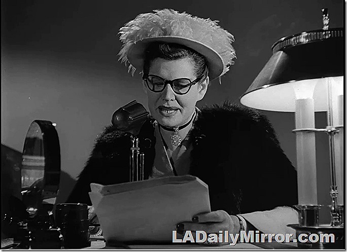 Woman in elaborate hat speaking into microphone. She wears a fur coat. 