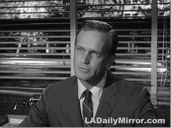 Man in coat and tie, venetian blinds in the background. 