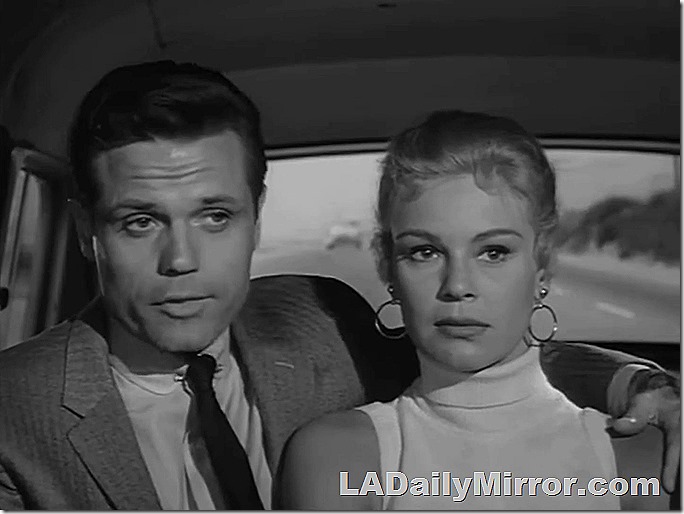 Man and woman riding in car. the man has his arm around the woman. He wears a coat and tie. She has big earrings. 