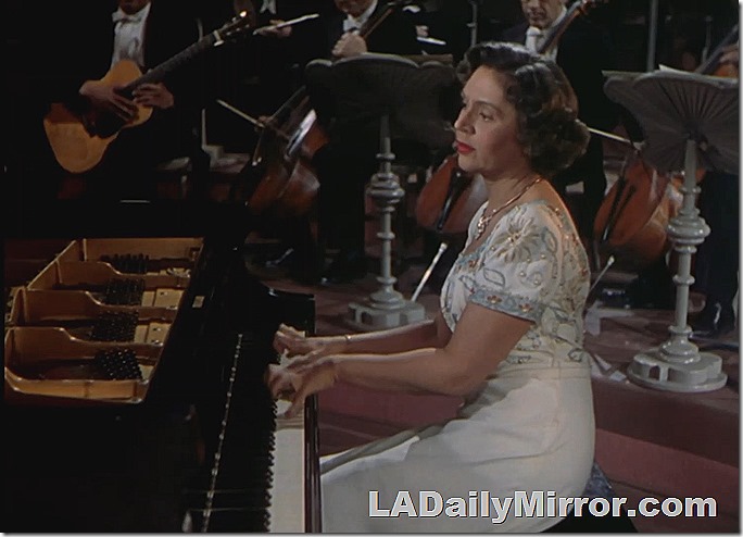 Woman at a grand piano, orchestra in the background. 