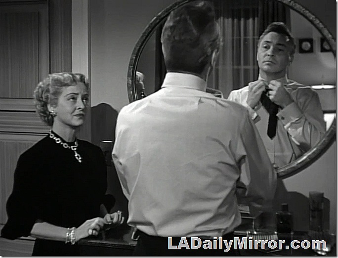 Man facing a mirror to tie his tie. A woman watches him. 