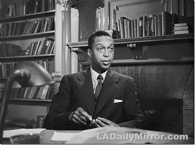 Man in suit, sitting at desk. Behind him are books and a fireplace. Man in suit, sitting at desk. Behind him are books and a fireplace.
