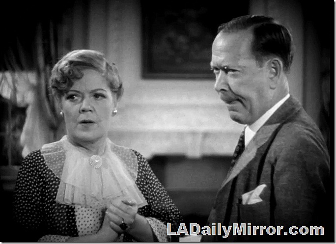 Woman in high-necked dress with decorative ruffle. Man in suit. Both have concerned expressions. 