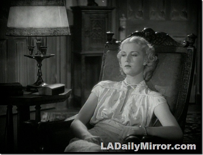 Woman sitting in heavy, ornate chair, wearing a fashionable outfit. Her eyes are downcast and she has a pensive look. 
