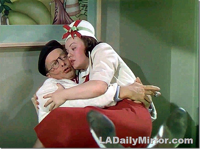 Mystery woman sitting in mysterious man's lap. He is wearing a beret and glasses. She is wearing a skirt, blouse and fancy hat. 