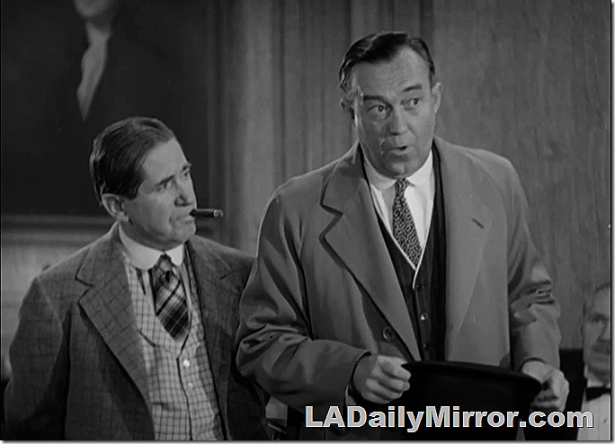 Two men. First is wearing loud vest, old-fashioned collar and is smoking a cigar. The second is tall, wearing an overcoat, vest and tie. 