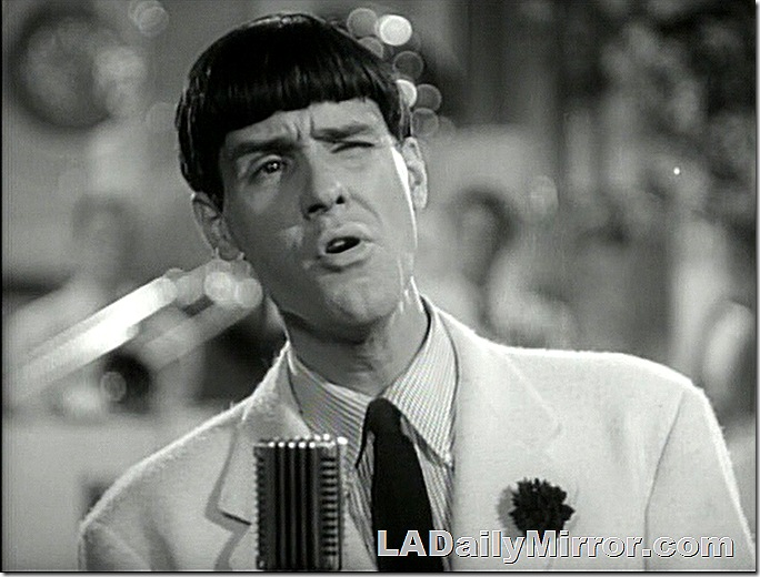 Man at microphone. He's wearing a coat and tie with a dark flower in his lapel. 