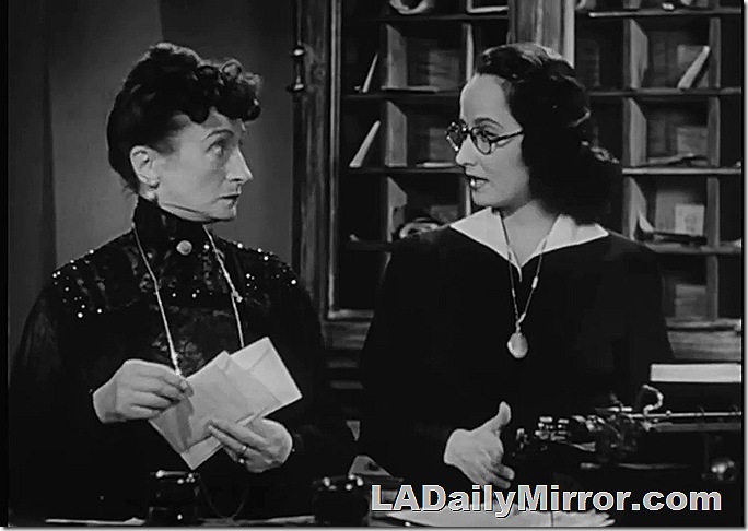 Woman in high collar dress looking at younger woman wearing glasses. 