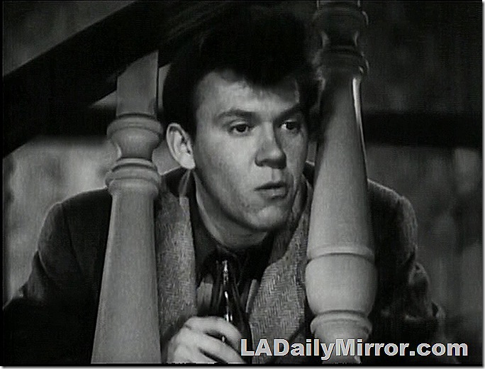 Man holding pop bottle, looking through stair railing. 