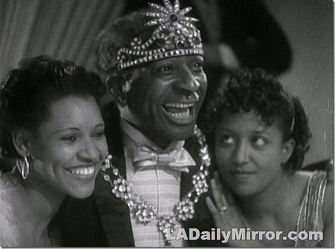 Man in tuxedo with jeweled headpiece and necklace, with two lady companions. 