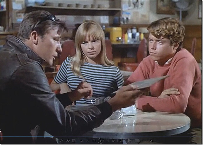 Two men and a woman at a diner. The man in a leather jacket is looking at a menu. The woman in the striped top and the youth in the sweater are looking at him. 